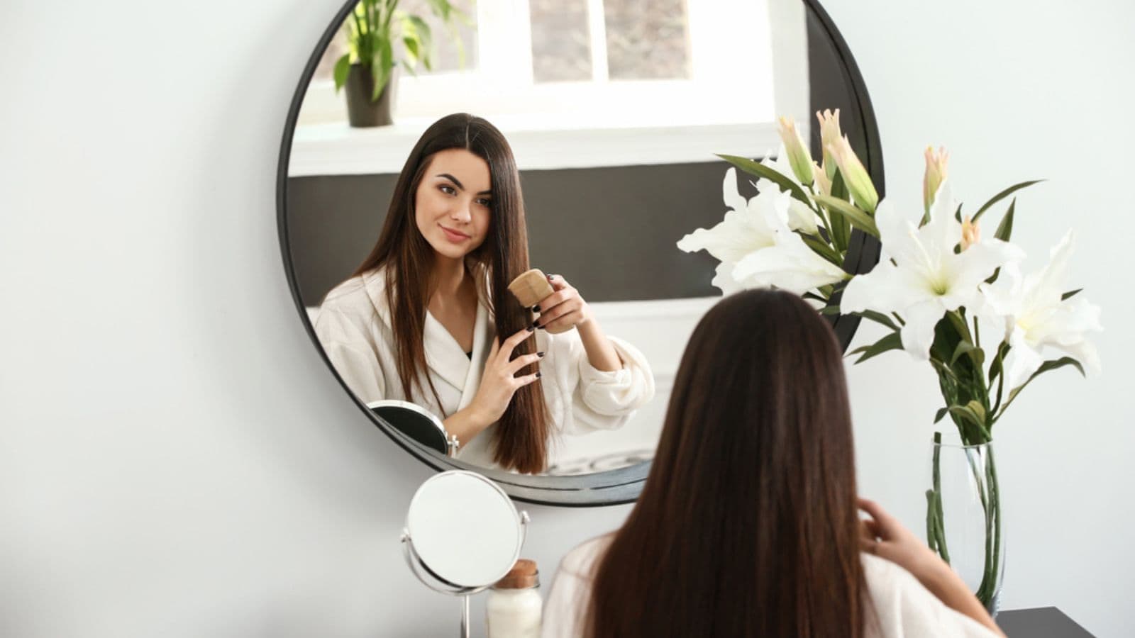 Beautiful young woman combing her healthy long hair near mirror at home
