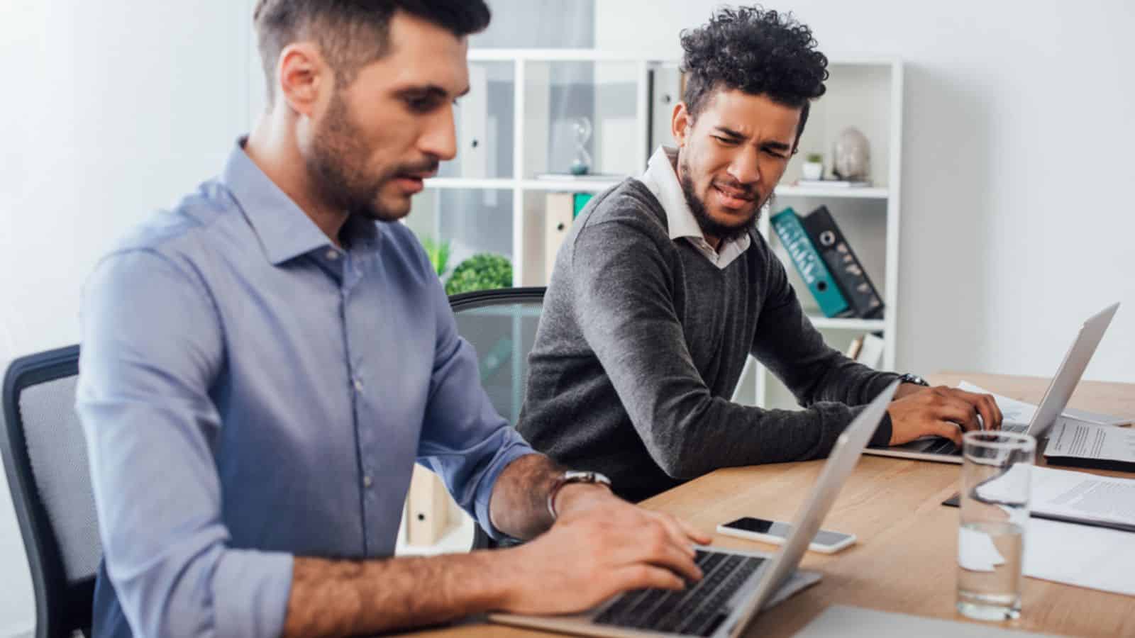 Confused african american businessman looking at colleague using laptop