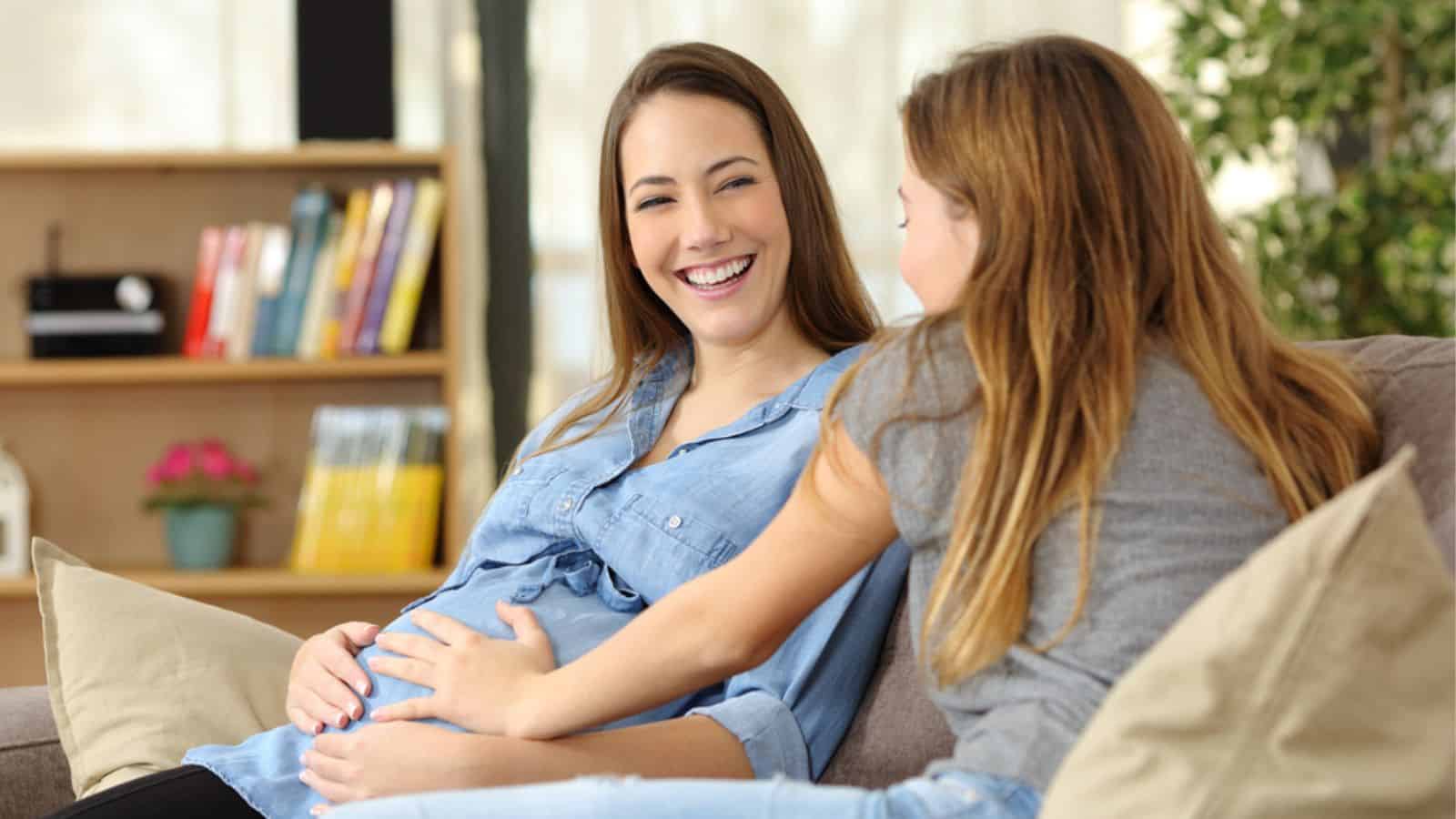 Laughing pregnant woman talking with a friend who touches her belly sitting on a couch
