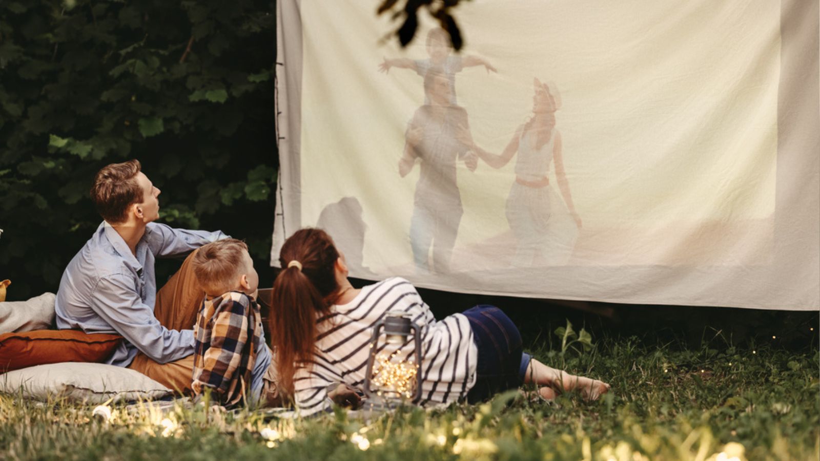 Parents and son sitting on blanket with pillows and watching movie projected on cloth in summer evening in yard