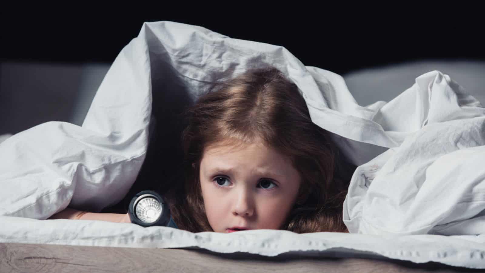 scared child hiding under blanket and holding flashlight