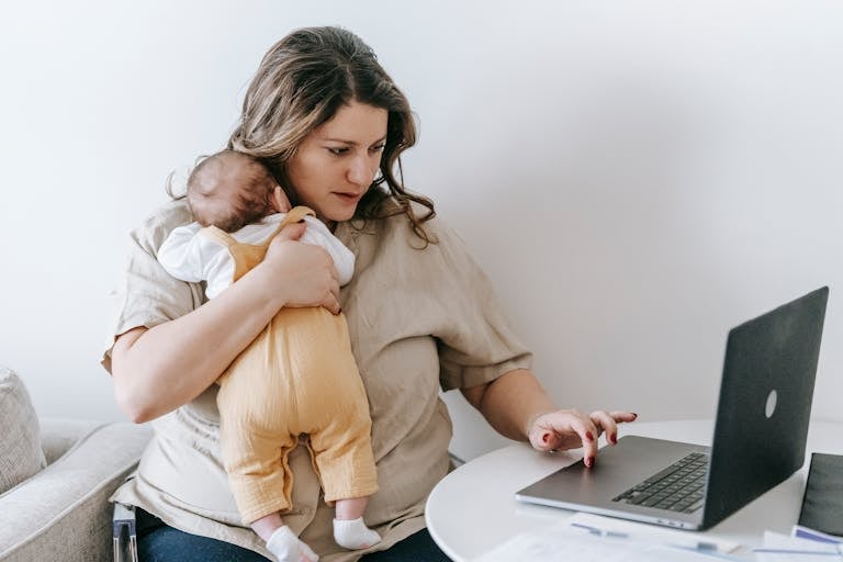 Concentrated young female freelancer embracing newborn while sitting at table and working remotely on laptop at home; pumping schedule