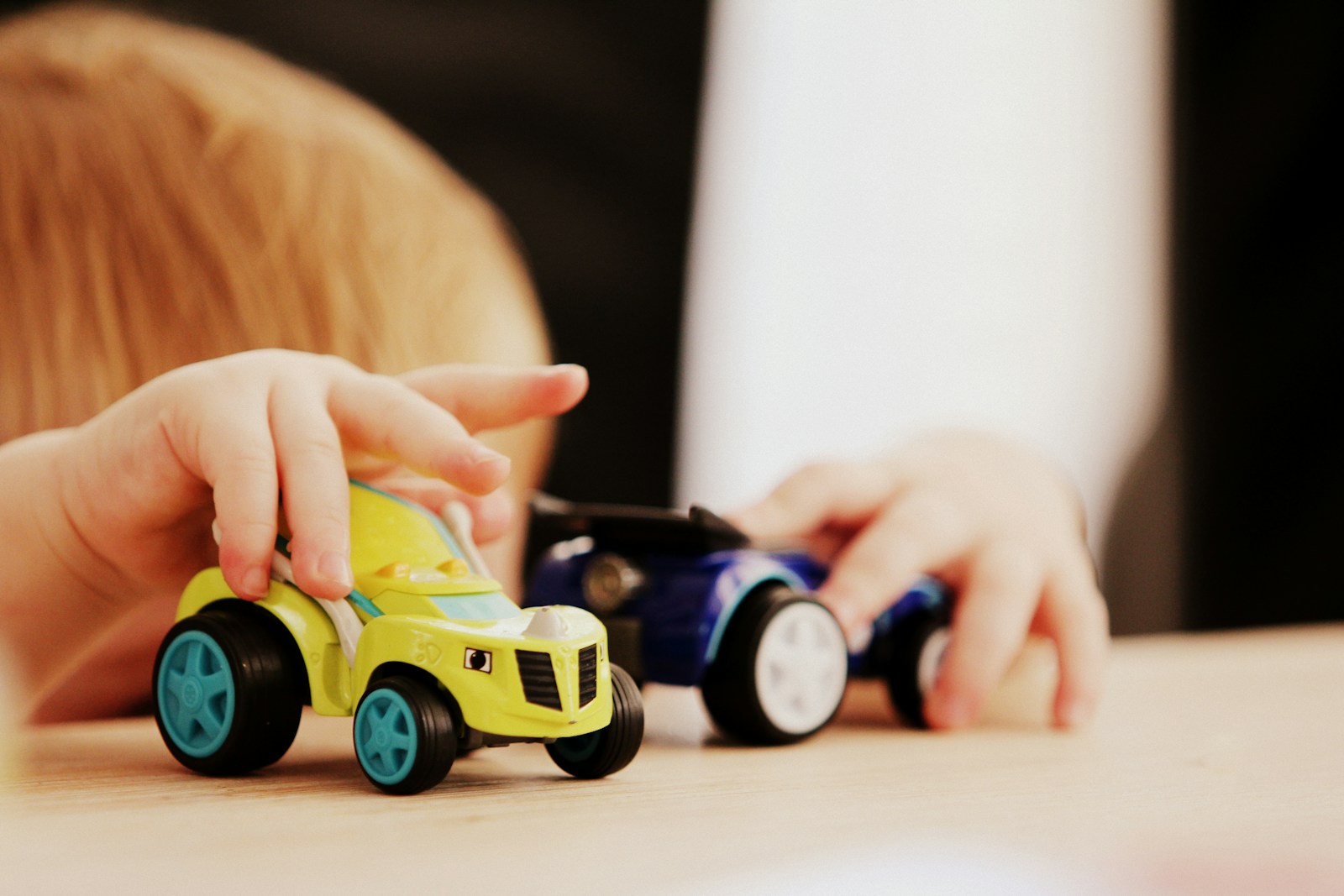 child playing with two assorted-color car plastic toys on brown wooden table; object permanence