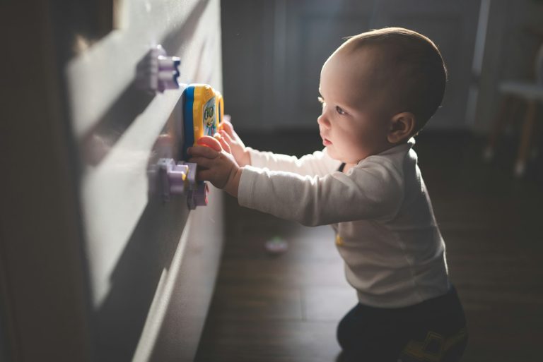 baby in white long sleeve shirt and black pants holding blue plastic toy; childproof