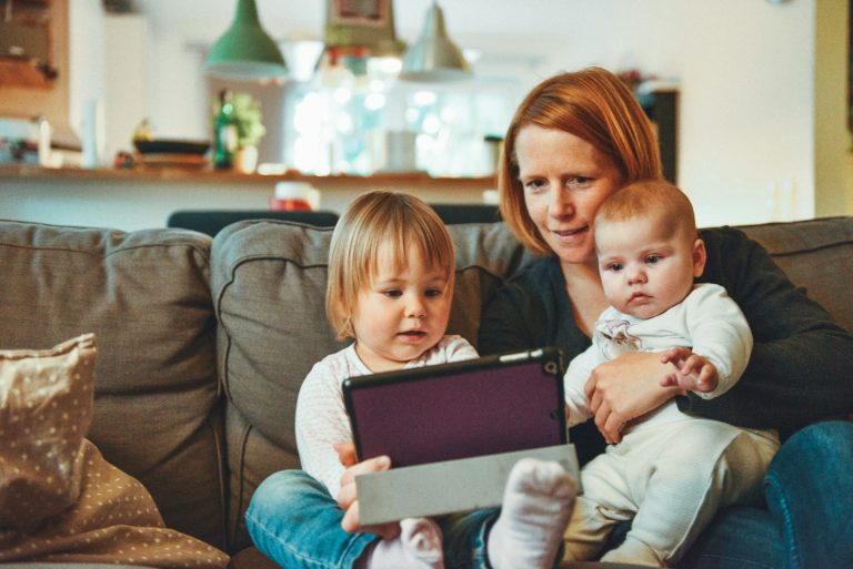 two babies and woman sitting on sofa while holding baby and watching on tablet; parenting trends