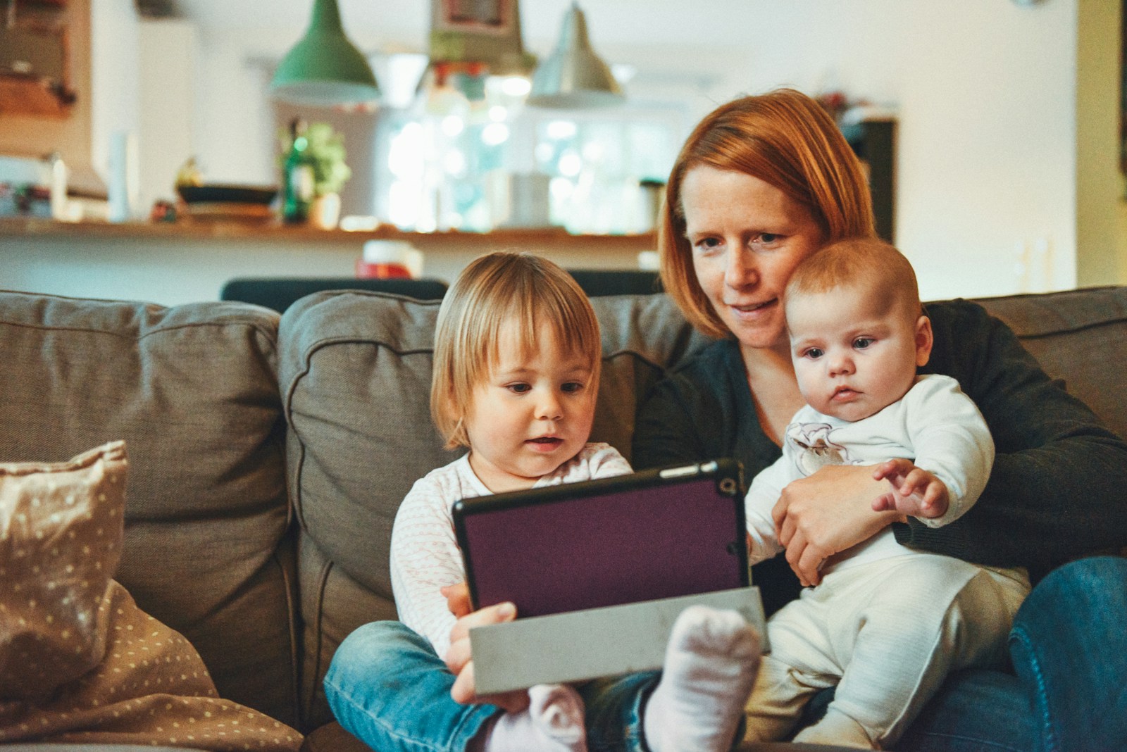 two babies and woman sitting on sofa while holding baby and watching on tablet; parenting trends