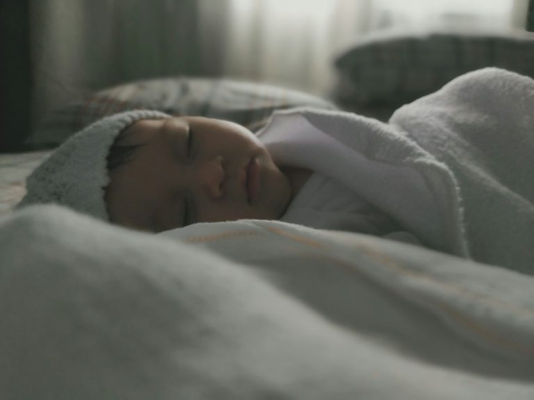 woman in white blanket lying on bed; co-sleeping vs. crib